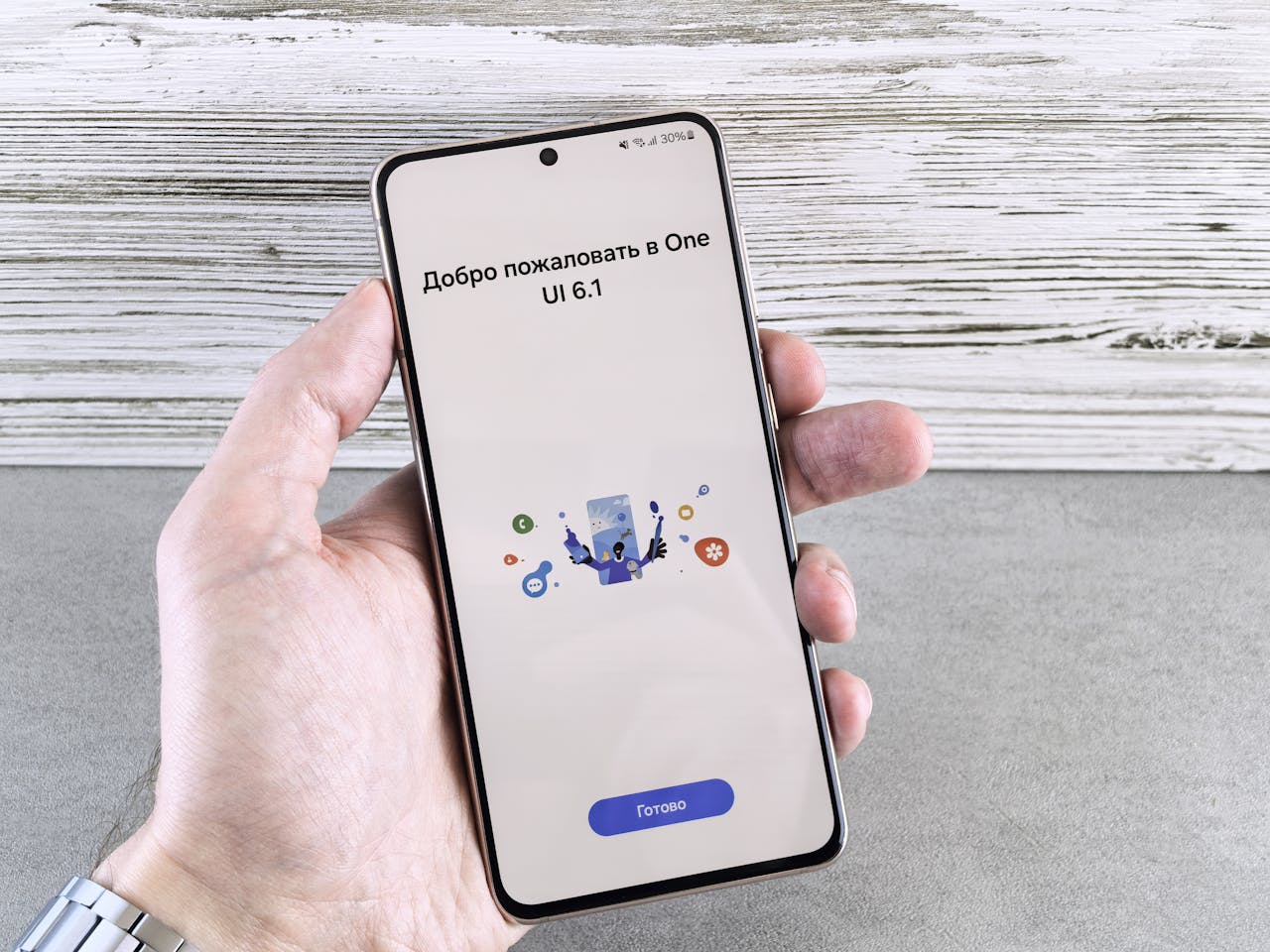 A close-up of a hand holding a smartphone displaying the One UI 6.1 welcome screen indoors.